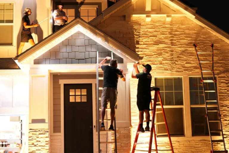 Installers on ladders and the roof mounting permanent lights along a home’s front gable at night