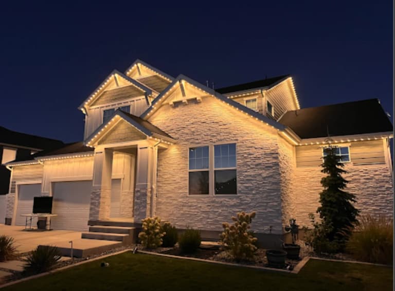 Two-story home at night with warm white permanent lights outlining every gable and roofline